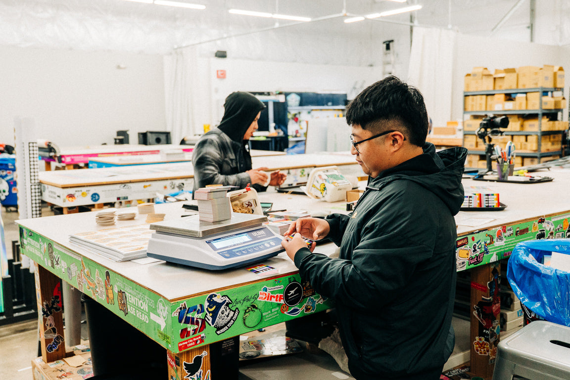 People working at a table in a warehouse setting with various items and equipment around.