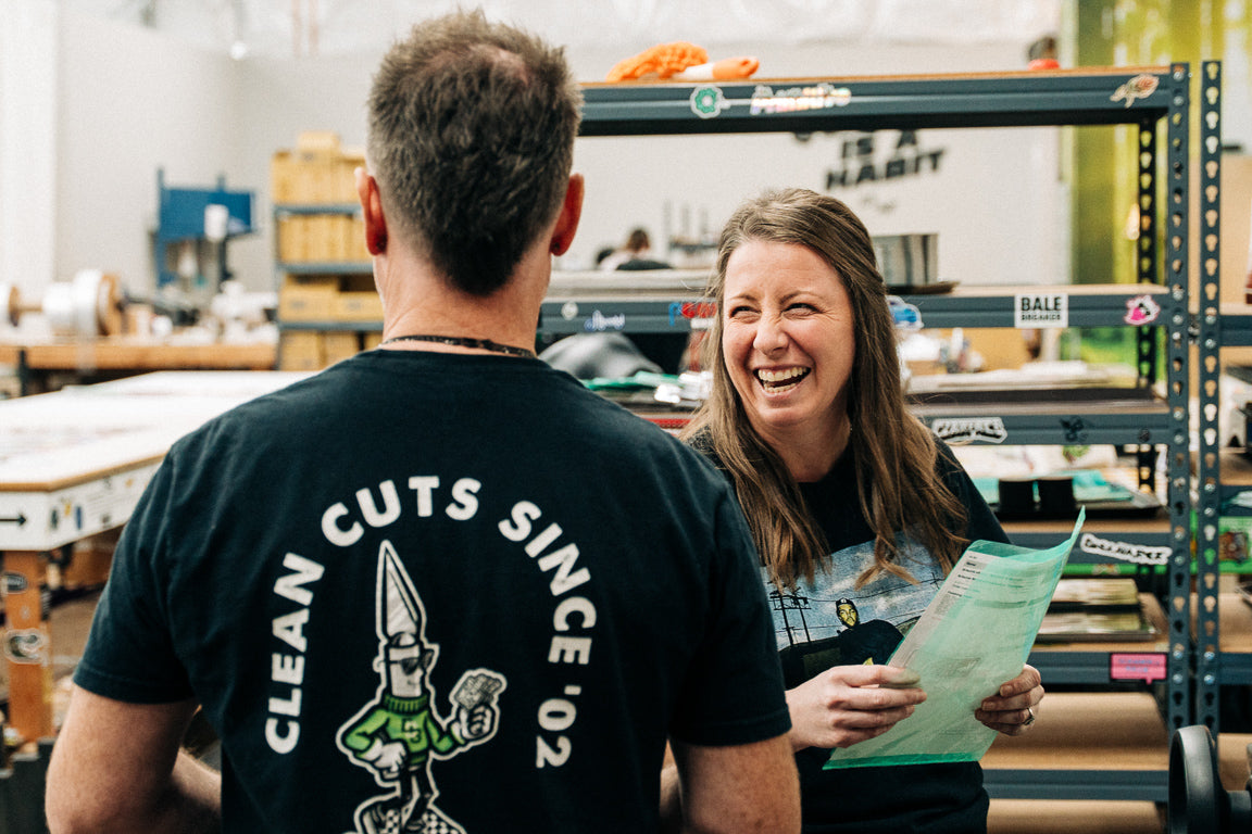Two people in a warehouse setting, one wearing a 'Clean Cuts' t-shirt, with shelves and products in the background.