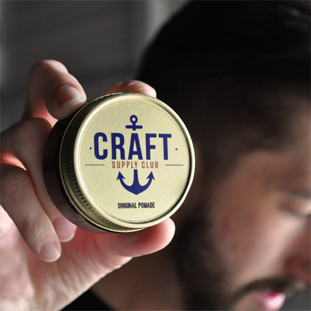 Hand holding a jar of 'Craft Supply Club' pomade with a blurred background