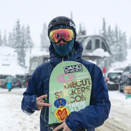 Person holding a snowboard with visible branding in a snowy outdoor setting